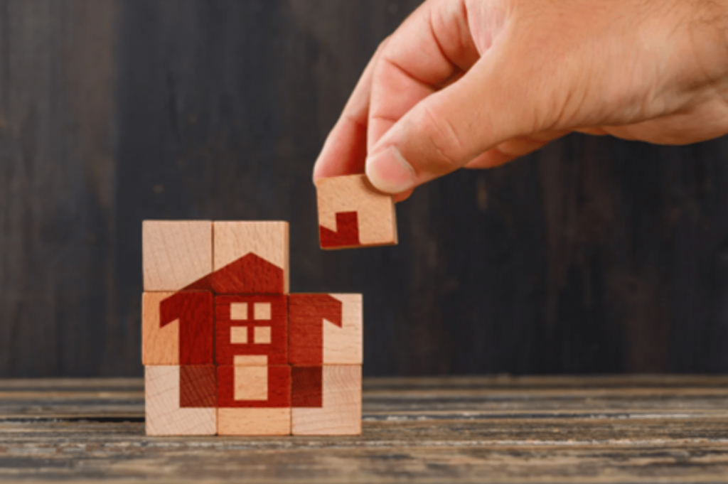 Main plaçant une pièce de puzzle sur une structure en bois ressemblant à une maison, sur un fond en bois.